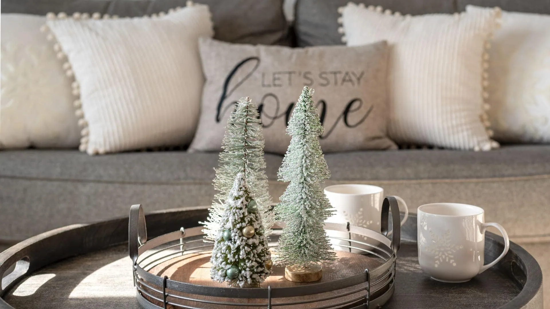 Minimalist Christmas decor with "Let's Stay Home" pillow and small decorative trees on a tray.